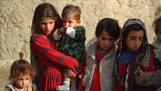 A group of children in colorful, traditional clothing stands in front of a loamy wall.