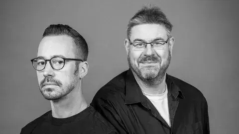 Die beiden Musiker sitzen nebeneinander für ein Portrait. Das Foto ist in Schwarz-Weiß.