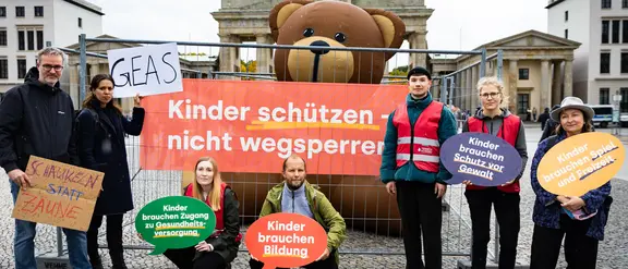 Eine Gruppe von Menschen mit Schildern und einem Riesenteddy steht vor dem Brandenburger Tor
