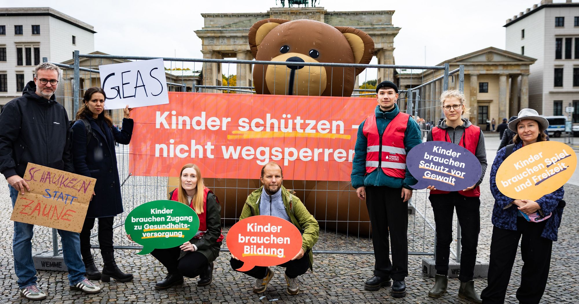 Eine Gruppe von Menschen mit Schildern und einem Riesenteddy steht vor dem Brandenburger Tor