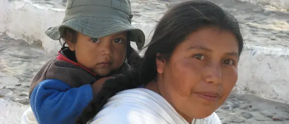 In Guatemala trägt eine Mutter ihr Kind auf dem Rücken, während sie auf einer steinernen Treppe sitzt. Die Szene zeigt die enge Bindung und traditionelle Lebensweise in der Region.