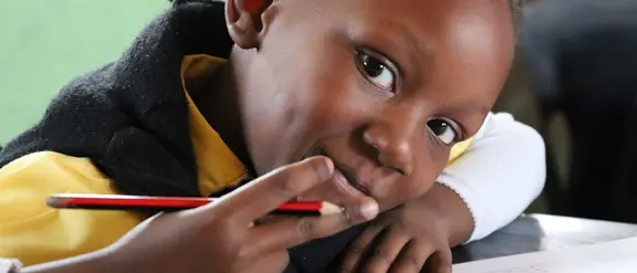 Ein Kind aus Südafrika zeigt Peace mit seiner rechten Hand, in der es auch einen Bleistift hält. Das Kind lehnt auf einem Tisch mit einem Schulheft darauf.