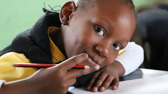 Ein Kind aus Südafrika zeigt Peace mit seiner rechten Hand, in der es auch einen Bleistift hält. Das Kind lehnt auf einem Tisch mit einem Schulheft darauf.