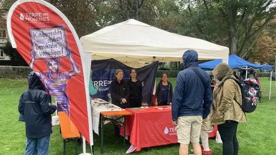 El puesto de información rojo de la organización de derechos de la infancia Terre des Hommes destaca sobre un prado verde. Tres personas detrás del puesto conversan con personas interesadas que se encuentran frente a él.