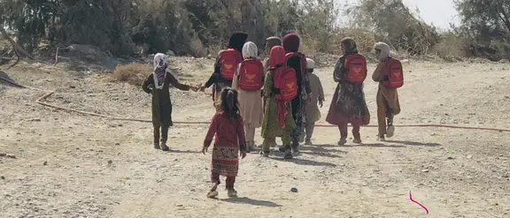 Kinder mit roten Rucksäcken gehen einen staubigen Weg entlang in einer ländlichen Gegend auf ihrem Schulweg.