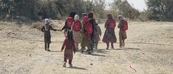 Los niños con mochilas rojas van por un camino polvoriento a lo largo de una zona rural en su camino a la escuela.