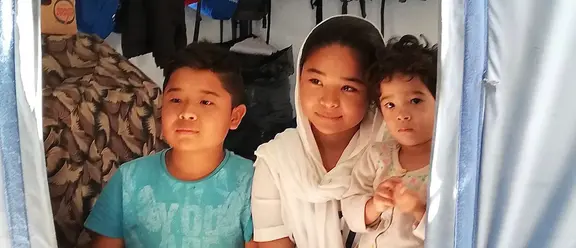 Drei Kinder sitzen im Eingang eines Zeltes in einem Flüchtlingslager in Griechenland, vereint in ihrem Zusammenhalt.