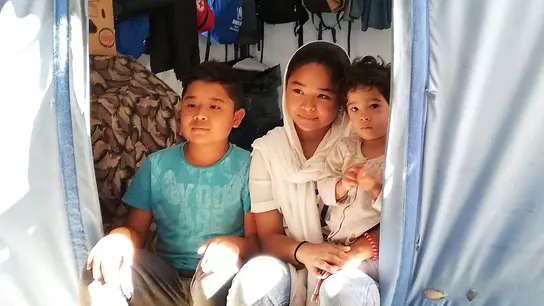 Tres niños se sientan en la entrada de una tienda de campaña en un campo de refugiados en Grecia, unidos en su cohesión.
