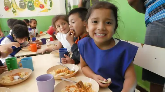  La niña sonriente se sienta en una cantina en El Salvador con otros niños y come una comida.
