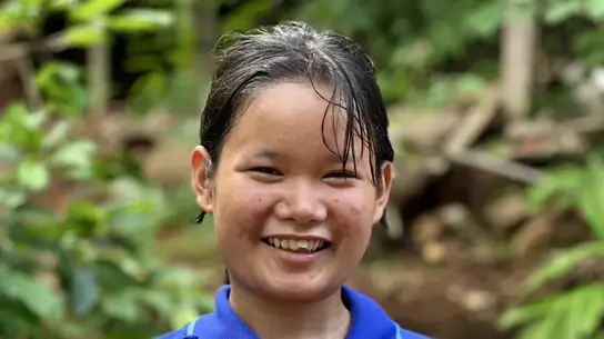 Ein junges Mädchen aus Myanmar namens Melada steht vor einem grünen Wald und lächelt für das Portraitbild. Sie trägt ein blaues Polo-T-Shirt.