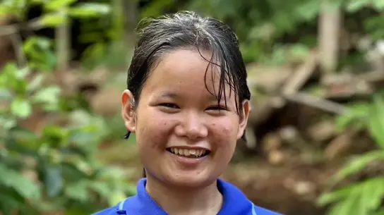 Ein junges Mädchen aus Myanmar namens Melada steht vor einem grünen Wald und lächelt für das Portraitbild. Sie trägt ein blaues Polo-T-Shirt.