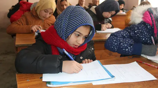 Junge Schülerinnen in warmer Winterkleidung sitzen in einem Klassenzimmer und schreiben konzentriert Notizen. Sie tragen bunte Schals und Mützen.