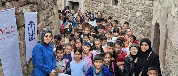 Zwischen alten Stadtmauern steht eine große Gruppe von Kindern in der Türkei, die eine Wanderung machen. Betreuerinnen stehen daneben und sie halten Plakate und Banner in der Hand.