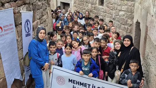 Zwischen alten Stadtmauern steht eine große Gruppe von Kindern in der Türkei, die eine Wanderung machen. Betreuerinnen stehen daneben und sie halten Plakate und Banner in der Hand.